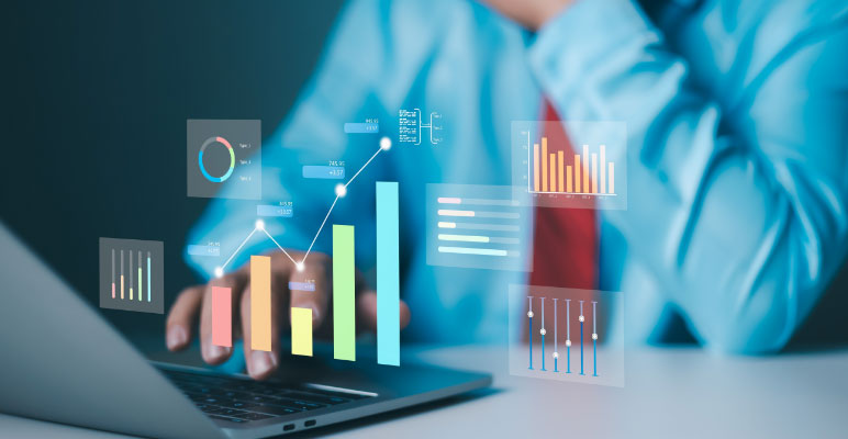 Photograph of man standing in front of three people sitting at desks with laptops while he is pointing at a bar graph on a large screen.